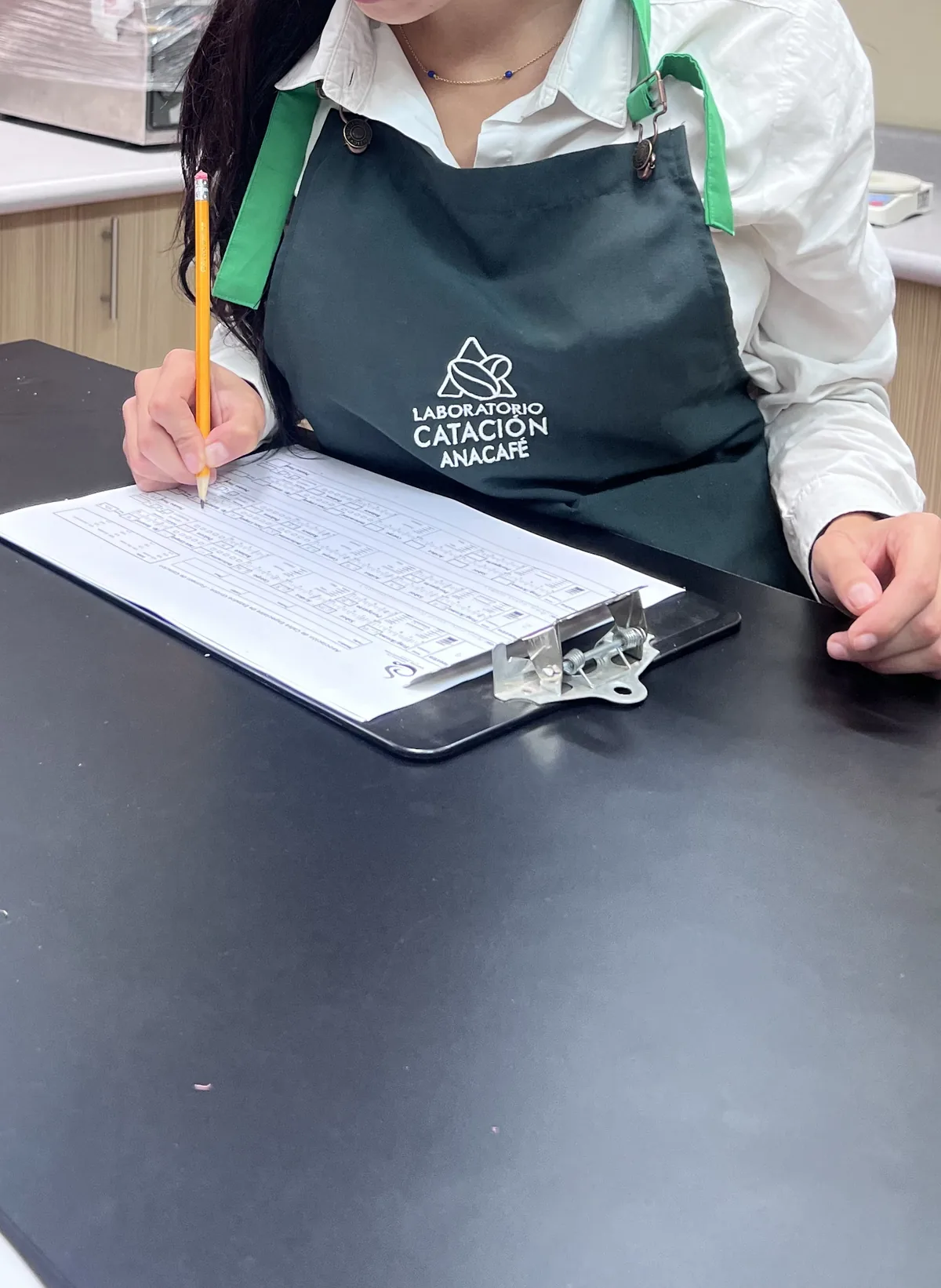 A woman in a green Anacafé cupping laboratory apron writes on a clipboard at a lab table.